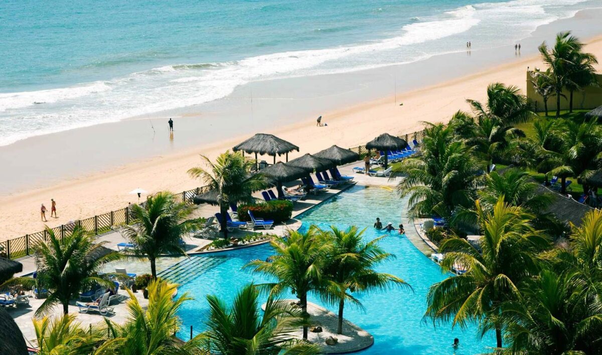 Piscina del resort frente a la playa en Brasil, rodeada de palmeras y sombrillas de paja.
