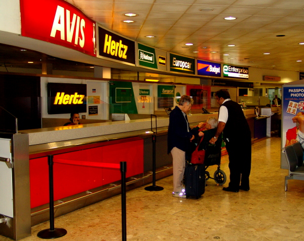 El alquiler de autos en el aeropuerto de Miami es una buena opción. Averigua precios en las agencias como Avis, Hertz, Budget y Enterprise. Mostrador de alquiler de autos en un aeropuerto, con logos de compañías como Avis, Hertz, Europcar y Budget. Dos personas en el mostrador reciben asistencia.