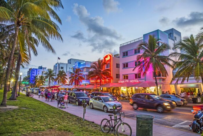 Vista de Ocean Drive en Miami Beach al atardecer, con luces de neón en los edificios, palmeras a los lados y autos circulando por la calle.