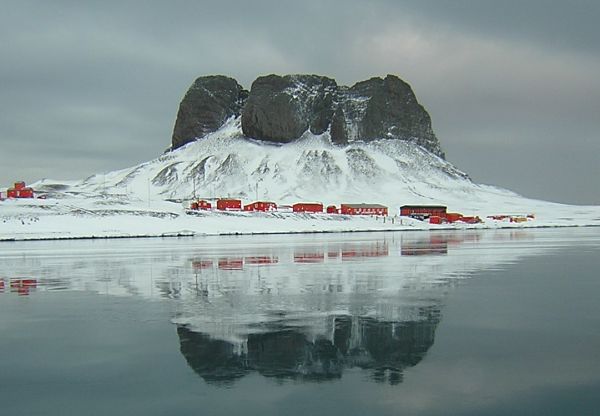 Las bases científicas: la vida en el continente Blanco Base antártica de investigación con edificios rojos bajo una formación rocosa y nevada. La montaña y los edificios se reflejan en el agua helada.