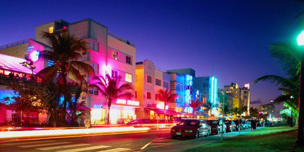 Vista nocturna de Ocean Drive en Miami Beach, con edificios art déco iluminados por luces de neón y autos estacionados en la calle. La escena captura la vibrante vida nocturna del área.