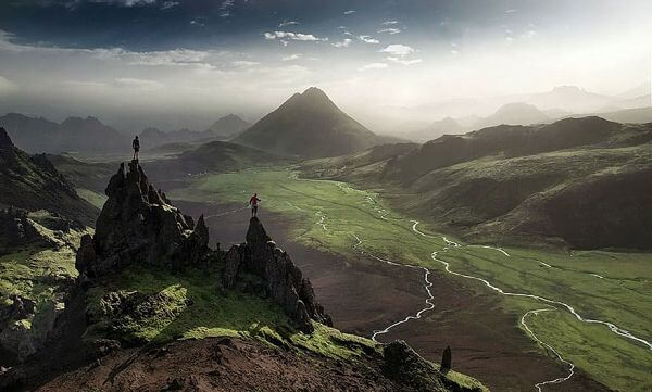 Senderismo en montañas y valles de Islandia Paisaje montañoso de Islandia con senderistas en lo alto de un pico rocoso y un valle verde atravesado por ríos.