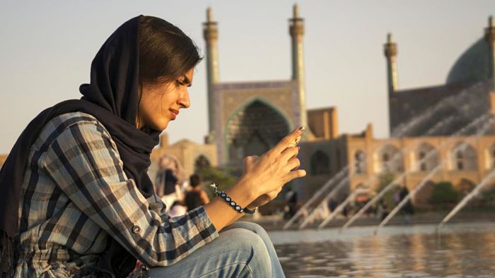 Mujer joven con pañuelo en la cabeza utilizando su teléfono móvil frente a una mezquita islámica con cúpula y minaretes