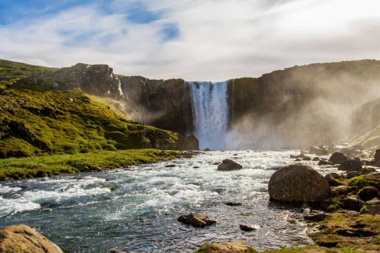 Cascada rodeada de campos verdes y un r&iacute;o en Islandia durante los meses de clima m&aacute;s templado.